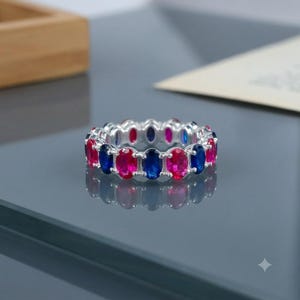 以下が含まれることがあります： 鮮やかなブルーと明るいピンクのオーバルカットの宝石が交互に並び、磨かれた金属製の枠にセットされたシルバーのエタニティリング。対称的でカラフルなデザインが滑らかな表面に映り込んでいます。