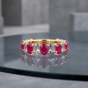 Könnte beinhalten: Ein goldener Eternity-Ring mit einem sich wiederholenden Muster aus oval geschliffenen Edelsteinen in abwechselnden Farbtönen von leuchtendem Rot und zartem Lavendel. Die Steine sind in eine polierte Goldfassung eingefasst.