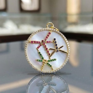May include: A circular gold pendant featuring a white mother-of-pearl inlay. The surface is decorated with three dragonfly motifs crafted from small red, blue, and green gemstones. The outer edge is lined with tiny clear pavé-set crystals.