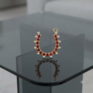 May include: A gold horseshoe-shaped pendant featuring a row of deep red circular stones along the inner curve and a series of small clear circular stones set along the outer edge, with a small circular jump ring attached at the top right side.