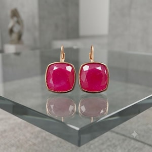 Op de afbeelding: Een paar oorbellen met klapbrisure en vierkant geslepen, gefacetteerde felroze edelstenen in een gepolijste goudkleurige metalen zetting. De oorbellen hebben een reflecterend oppervlak met duidelijk zichtbare spiegelbeelden eronder.