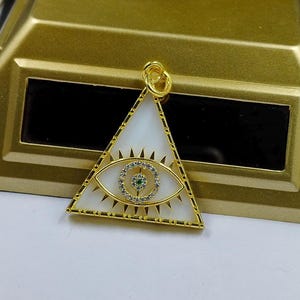 Op de afbeelding: Goudkleurige hanger in een driehoekige vorm, met een wit middenstuk met een oogontwerp. Het oog is gedetailleerd met een cirkel van heldere stenen en een groen centrum. De hanger heeft een lus aan de bovenkant voor een ketting.
