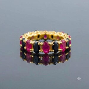 May include: A gold eternity band featuring an alternating pattern of oval-cut vibrant pink rubies and deep black onyx gemstones. The stones are set in a continuous circular arrangement, creating a striking contrast between the gold and the gems.