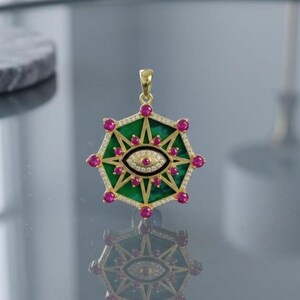 May include: A gold pendant featuring a central eye motif surrounded by a green inlay and a starburst pattern. The piece is accented with small round pink stones at each point and along the inner eye, with clear pavé stones lining the outer frame.