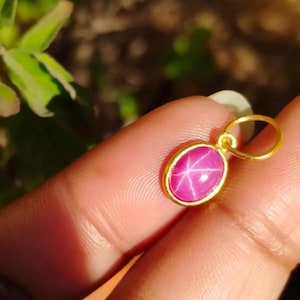 May include: A gold pendant with an oval, pink gemstone featuring a six-ray star pattern. The gemstone is set in a gold bezel and has a gold loop for a chain. The pendant is held up by a finger.