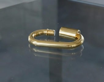 Può includere: Un moschettone in metallo dorato di forma ovale, caratterizzato da una finitura liscia e lucida e da un meccanismo di chiusura a vite su un lato. Il moschettone è aperto, rivelando la filettatura interna sull'estremità del connettore.