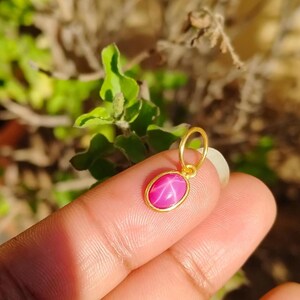 May include: A gold pendant with a pink oval-shaped gemstone. The gemstone has a star-like pattern. The pendant is held between fingers against a blurred green and brown background.