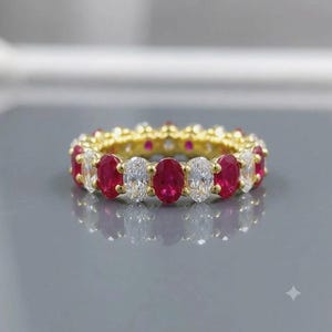 May include: A gold eternity band featuring an alternating pattern of oval-cut red rubies and clear white diamonds. The gemstones are set in a continuous row around the circular band, creating a vibrant contrast between the deep red and clear stones.