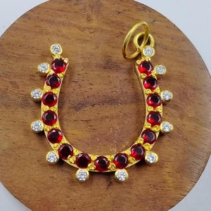 May include: A gold horseshoe pendant featuring a series of round, deep red gemstones set along the inner curve, accented by smaller, clear, round white stones placed at intervals along the outer edge, complete with a gold circular bail at the top.
