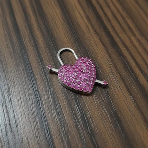 May include: A silver heart-shaped padlock pendant with a pink gemstone surface pierced by a silver arrow. The padlock's shackle is curved. The pendant is on a wooden surface.