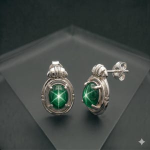 Puede incluir: Un par de pendientes de plata con piedras verdes ovaladas y un patrón de estrella. Los pendientes tienen un diseño detallado y ornamentado alrededor de la gema, con un cierre de poste y mariposa.