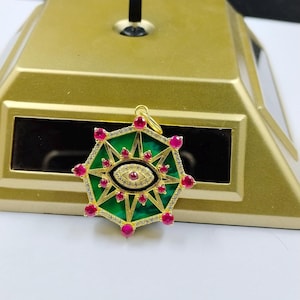 May include: Gold pendant featuring a green enamel design with a central eye motif. The pendant is embellished with red gemstones and small clear crystals, forming a starburst pattern. It includes a loop for a chain.