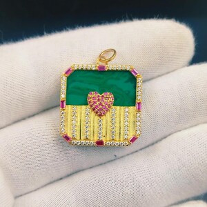 May include: A gold pendant with a rectangular shape, featuring a green center and a pink heart. The pendant is adorned with small, sparkling stones and pink rectangular accents. The design includes vertical gold stripes.