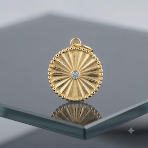 May include: A circular gold pendant featuring a sunburst pattern of radiating lines and a beaded outer edge. A single small, clear, round-cut gemstone is set in the centre of the pendant. A small gold jump ring is attached to the top for hanging.