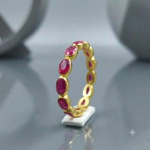 Könnte beinhalten: Ein goldener Eternity-Ring mit einer Reihe von oval geschliffenen, tiefroten Rubinen in einer durchgehenden Fassung. Der Ring steht aufrecht auf einem kleinen weißen rechteckigen Sockel und präsentiert die leuchtend roten Edelsteine.