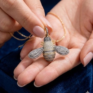 May include: A gold-toned necklace featuring a bee pendant. The bee pendant has a body with alternating bands of gold and black, and wings encrusted with small, sparkling stones. The necklace is held in a person's hands.