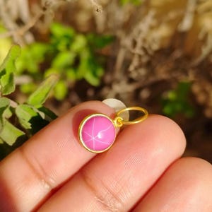 Può includere: Un pendente d'oro con una gemma rosa rotonda. La gemma ha un motivo a forma di stella sulla sua superficie. Il pendente è tenuto da un dito. Lo sfondo è sfocato con fogliame verde.