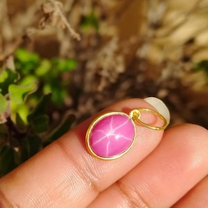 May include: A gold pendant with a pink oval-shaped gemstone. The gemstone has a star-like pattern across its surface. The pendant is held by a finger in front of a blurred green background.