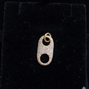 Può includere: Un ciondolo dorato a forma di chicco di caffè, incrostato di piccoli cristalli scintillanti. Il ciondolo ha un piccolo anello nella parte superiore per una catena e un'apertura circolare nella parte inferiore. Lo sfondo è un tessuto scuro e vellutato.