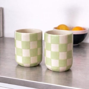 May include: Two cylindrical ceramic cups featuring a hand-painted light green and off-white checkered pattern. The cups have a smooth, rounded base and an open top, displaying a consistent grid design across the exterior surface of each vessel.