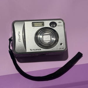 May include: Silver Fujifilm FinePix A345 digital camera with a black wrist strap. The camera has a lens, flash, and a 4.1-megapixel resolution. The camera is on a purple surface.