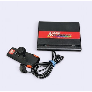 May include: Black Atari Flashback Classic Game Console with a red and white label. Includes a black joystick controller with an orange accent and a black cable. The console has power and reset buttons.