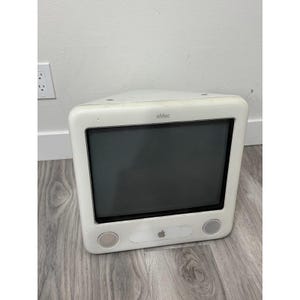 Puede incluir: Un ordenador de sobremesa Apple eMac blanco con una gran pantalla de tubo, un sistema de altavoces frontal a ambos lados de la base y el logotipo de Apple centrado bajo la pantalla. La unidad tiene una carcasa de plástico blanco redondeada.