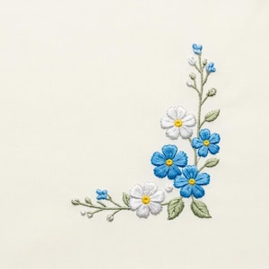 Könnte beinhalten: Gesticktes Blumendesign in der Ecke mit weißen und blauen Blüten, gelben Zentren und grünen Blättern auf cremefarbenem Stoff. Die Stickerei ist detailliert.