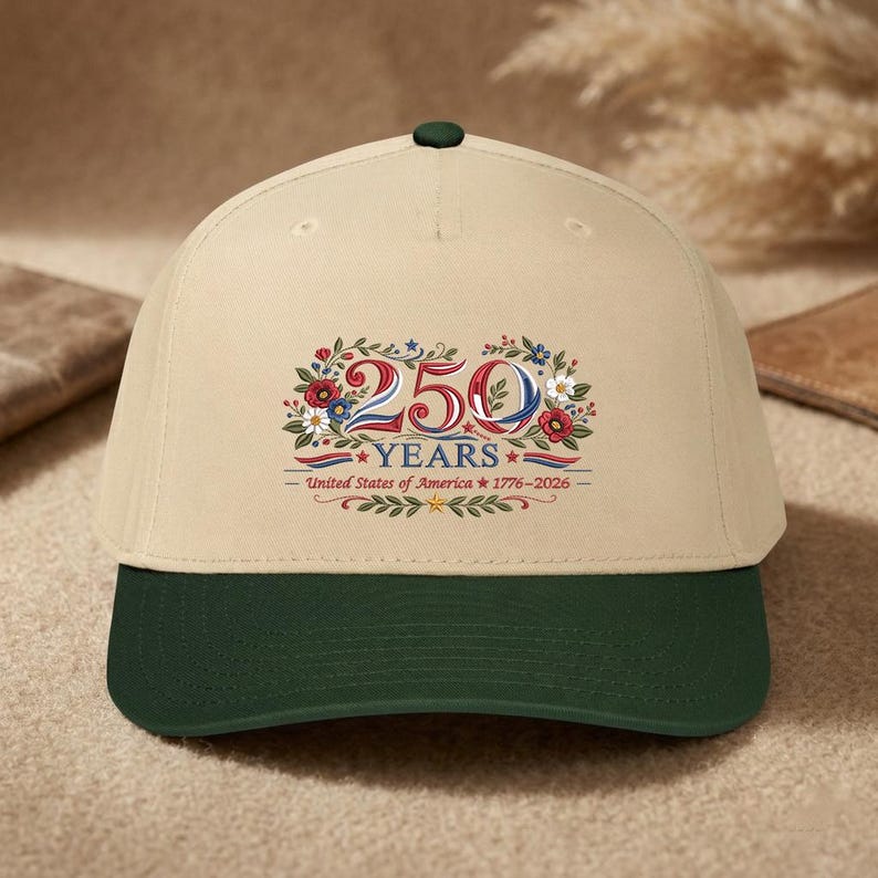 K&ouml;nnte beinhalten: Beige Baseballcap mit dunkelgr&uuml;nem Schirm und Knopf. Auf der Kappe befindet sich ein Blumendesign mit dem Text "250 YEARS - United States of America 1776-2026".