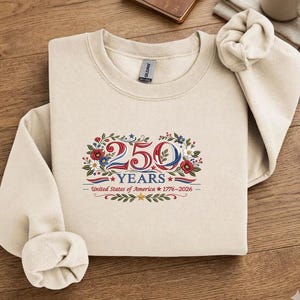K&ouml;nnte beinhalten: Ein cremefarbenes Sweatshirt mit Rundhalsausschnitt. Auf der Vorderseite befindet sich ein Blumendesign mit den Zahlen "250" in Rot und Blau, dem Wort "YEARS" und dem Text "United States of America 1776-2026".