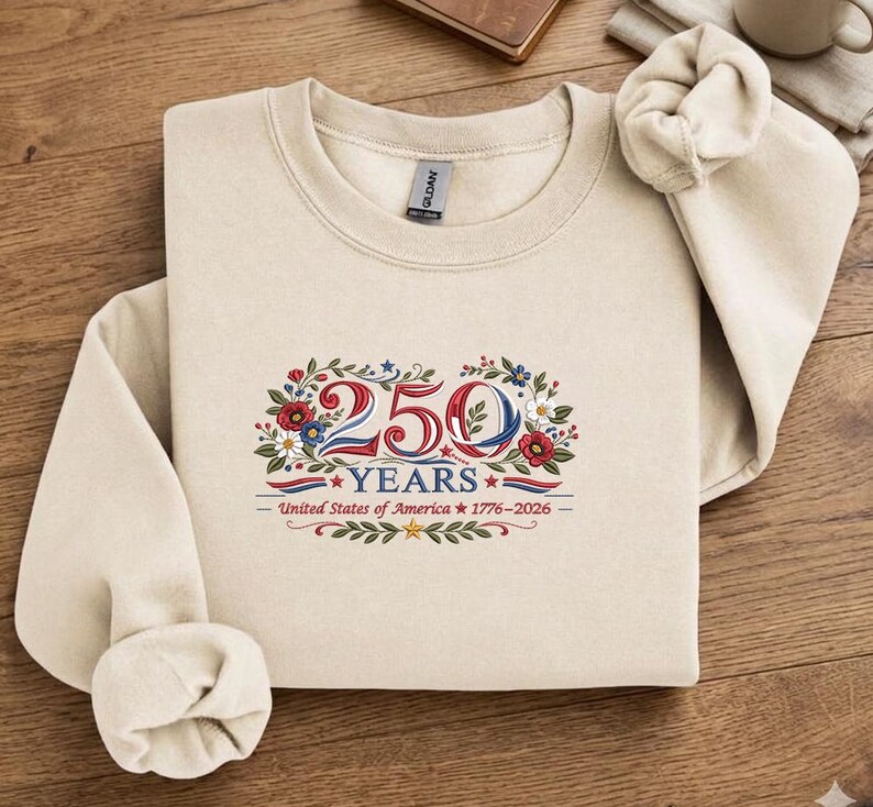 K&ouml;nnte beinhalten: Ein cremefarbenes Sweatshirt mit Rundhalsausschnitt. Auf der Vorderseite befindet sich ein Blumendesign mit den Zahlen "250" in Rot und Blau, dem Wort "YEARS" und dem Text "United States of America 1776-2026".