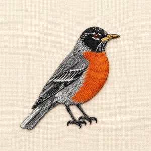 Op de afbeelding: Geborduurde patch van een roodborstje. De vogel heeft een zwarte kop, grijze vleugels en een feloranje borst. De patch is gedetailleerd met ingewikkelde steken, die de veren en de vorm van de vogel laten zien. De achtergrond is een neutraal beige.