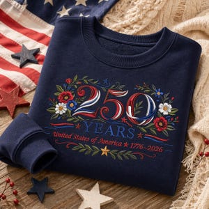K&ouml;nnte beinhalten: Marineblaues Sweatshirt mit gesticktem Blumendesign und dem Text "250 YEARS United States of America 1776-2026". Das Design zeigt rote, wei&szlig;e und blaue Farben, mit Sternen und floralen Akzenten. Das Sweatshirt ist gefaltet.