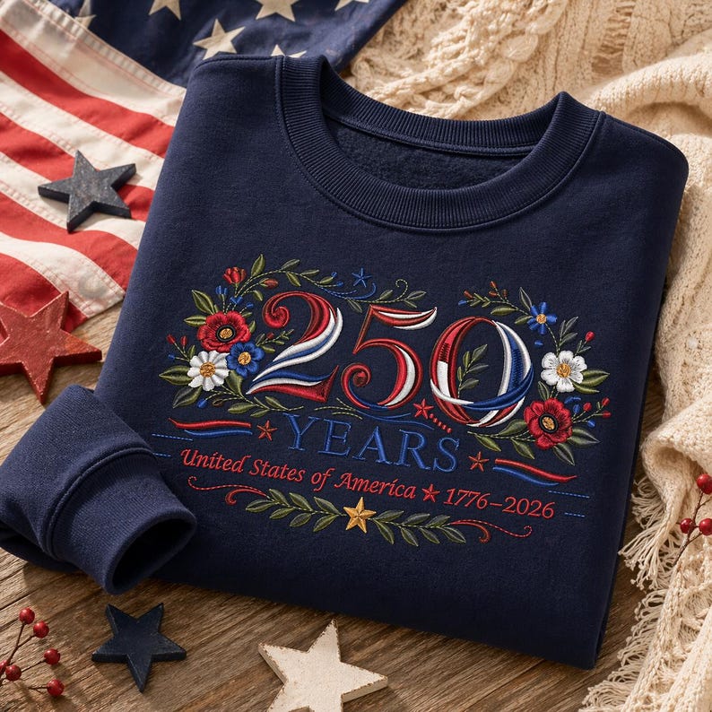 K&ouml;nnte beinhalten: Marineblaues Sweatshirt mit gesticktem Blumendesign und dem Text "250 YEARS United States of America 1776-2026". Das Design zeigt rote, wei&szlig;e und blaue Farben, mit Sternen und floralen Akzenten. Das Sweatshirt ist gefaltet.