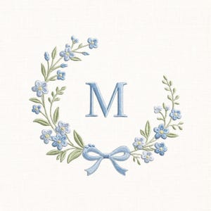 Könnte beinhalten: Gestickter Blumenkranz mit dem Buchstaben "M" in Hellblau. Der Kranz zeigt zarte blaue Blumen, grüne Blätter und eine passende Schleife unten. Das Design ist vor einem weißen Hintergrund platziert.