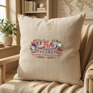 K&ouml;nnte beinhalten: Beiges quadratisches Kissen mit einem farbenfrohen Blumendesign und dem Text "250 YEARS - United States of America 1776-2026". Das Design weist rote, wei&szlig;e und blaue Elemente auf.