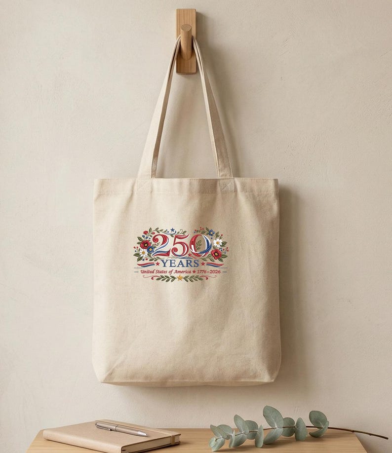 K&ouml;nnte beinhalten: Beige Tragetasche mit floralem Design und dem Text "250 YEARS United States of America 1776-2026" in Rot, Wei&szlig; und Blau. Die Tasche h&auml;ngt an einem Holzhaken.