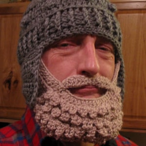 Può includere: Un cappello grigio all'uncinetto con una barba beige attaccata. La barba ha un design strutturato e arrotondato. La persona indossa una camicia a quadri rossa e blu. Il cappello copre la testa e le orecchie.