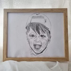 May include: A framed pencil sketch of a child with a baseball cap, smiling widely. The artwork is in a light wooden frame, set against a white backdrop. The sketch captures a moment of happiness.