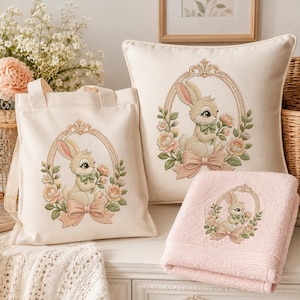 May include: A cream-colored canvas tote bag, a square throw pillow, and a folded light pink towel, each featuring an identical illustration of a cartoon rabbit inside an ornate oval frame surrounded by pink flowers and a ribbon bow.