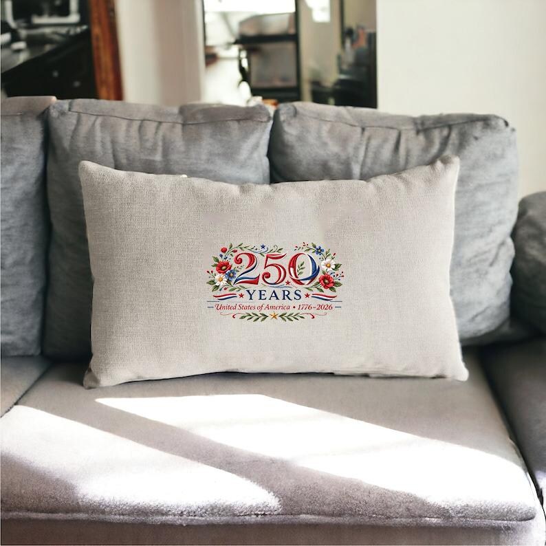 May include: A rectangular beige pillow with the text "250 YEARS" in red and blue, surrounded by floral and leaf accents. The text "United States of America 1776-2026" is below the main text. The pillow is on a gray couch.