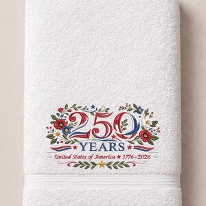 May include: White bath towel with an embroidered design celebrating 250 years of the United States of America. The design includes the numbers "250" in red, white, and blue, surrounded by floral and leaf elements.
