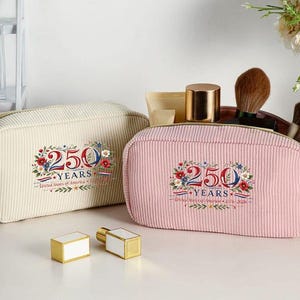 May include: Two corduroy cosmetic bags, one cream and one pink, with gold zippers. Each bag features a floral design with the text "250 YEARS United States of America 1776-2026". Two gold and white perfume bottles are in front.