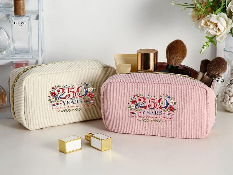 May include: Two corduroy cosmetic bags, one cream and one pink, with gold zippers. Each bag features a floral design with the text "250 YEARS United States of America 1776-2026". Two gold and white perfume bottles are in front.