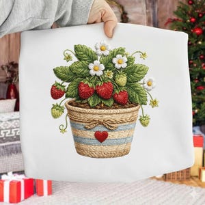 Puede incluir: Un cojín blanco con una ilustración detallada de una planta de fresa en una maceta tejida. La maceta tiene rayas azules y un corazón rojo. La planta tiene fresas rojas, hojas verdes y flores blancas.