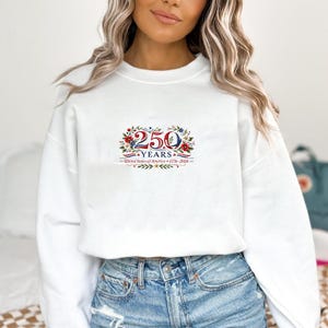 May include: White sweatshirt with a floral and text design. The design features the number "250" with the word "YEARS" below it, and the text "United States of America 1776-2026". The design is in red, white, and blue.