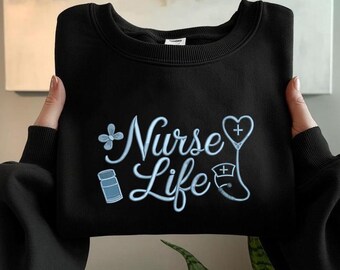 May include: Black sweatshirt with the words "Nurse Life" in light blue script. The design includes a flower, pill bottle, stethoscope, and cross symbols. The sweatshirt is held by hands.