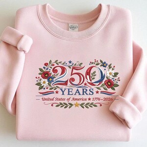 May include: A light pink sweatshirt with a crew neck and rolled-up sleeves. The front features an embroidered design with the text "250 YEARS" and "United States of America 1776-2026" in red, white, and blue, surrounded by floral elements.