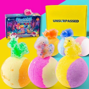 Könnte beinhalten: Eine bunte Auswahl an Badekugeln mit matschigen Tierfiguren. Die Badekugeln sind rund und haben verschiedene Farben, darunter Gelb, Rosa und Lila. Eine Schachtel mit der Aufschrift "Squishie Surprise" und eine gelbe Schachtel mit "UNSURPASSED" sind ebenfalls zu sehen.