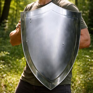 May include: A polished silver shield with a pointed base and curved top, held by a person. The shield's surface reflects light, and it is secured with rivets. The person is wearing a green t-shirt and dark trousers.